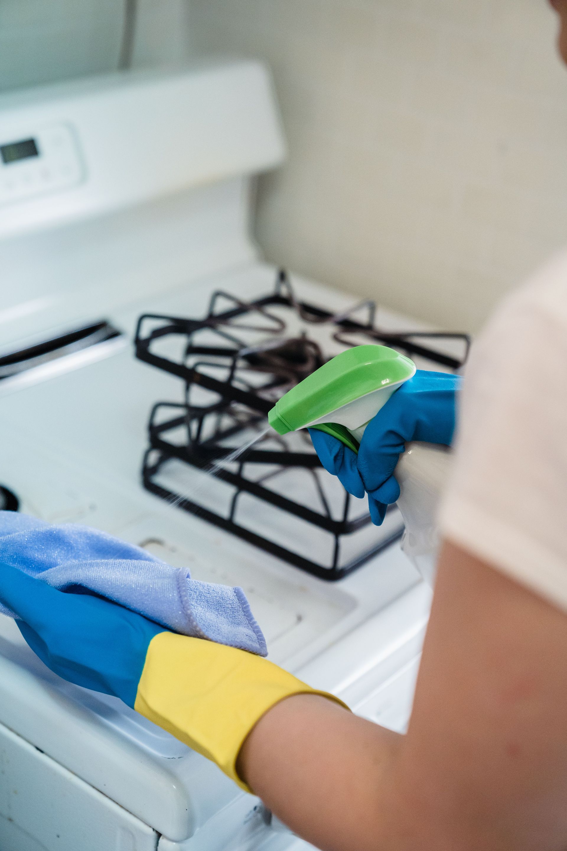 A person wearing gloves uses a spray bottle and cloth to clean a kitchen stove.